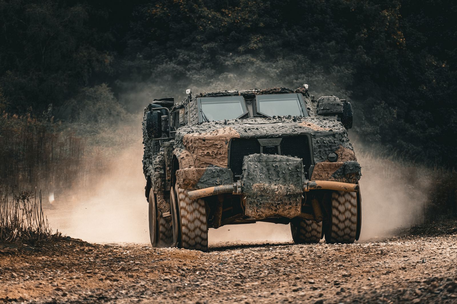 Camouflaged protected mobility vehicle kicking up dust on a gravel track